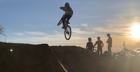 Merric Bayless 24 hits a jump overlooking the sunset at a local spot called the V. Many mountain bikers watch him while socializing with each other, providing a friendly atmosphere in the mountain biking community.