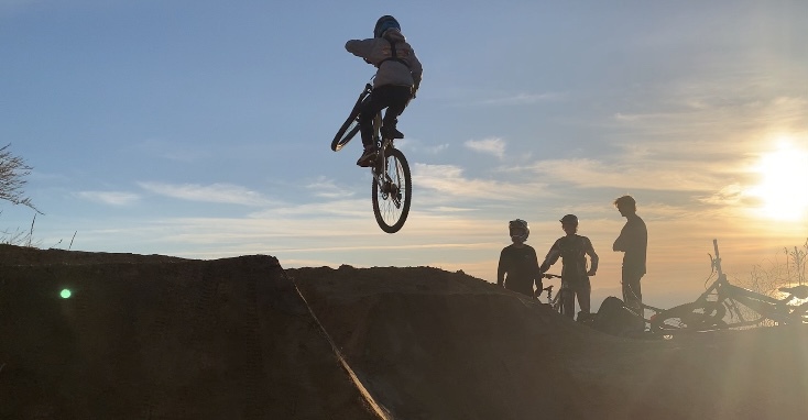Merric Bayless 24 hits a jump overlooking the sunset at a local spot called the V. Many mountain bikers watch him while socializing with each other, providing a friendly atmosphere in the mountain biking community.