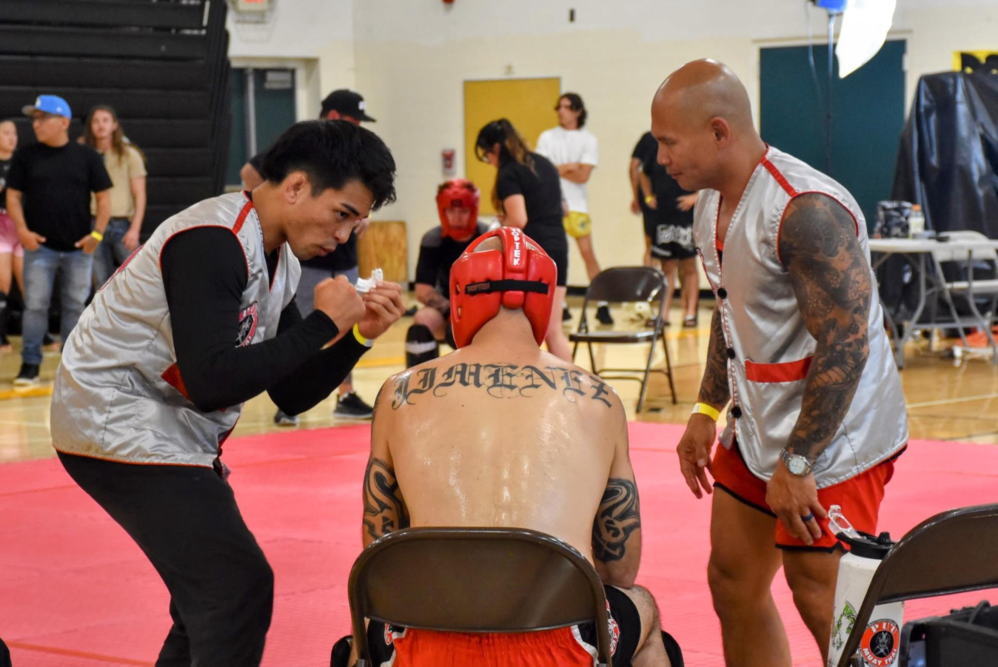 At the end of a second-round fight, a coach pictured on the left shadowboxes to demonstrate strategy while a second coach readies water for his fighter, who sits sweating at the edge of the mat. The scene unfolded at Eat, Sleep Teep, a Muay Thai event held at Ventura High School (Ventura), where coaching corners lined the competition mats and athletes and spectators packed the gym throughout the warm day.